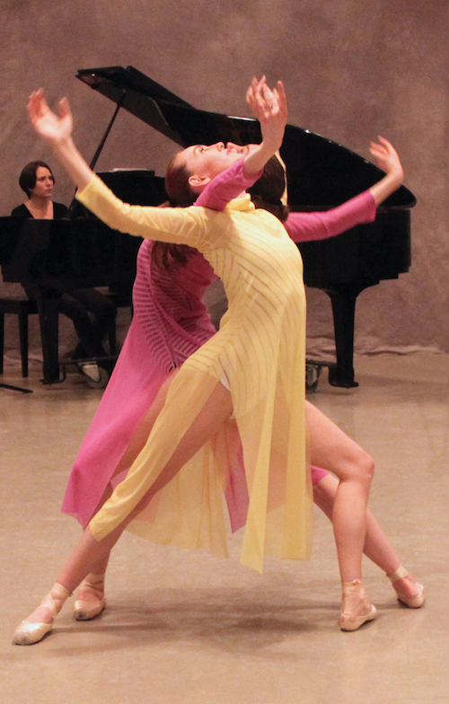 Two women lunge and arch their backs. Their faces look up to ceiling. A woman playing the piano is in the background.